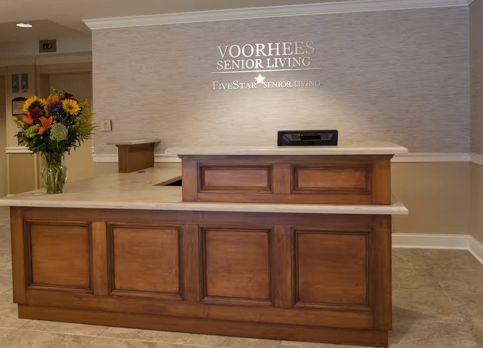 Lobby reception desk at Voorhees Senior Living with a wooden counter, wall sign, and a vase of flowers.