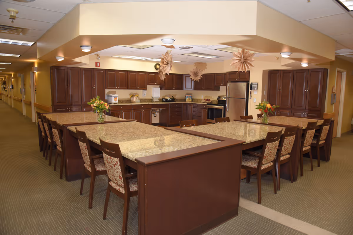 A spacious kitchen area with multiple granite-topped islands surrounded by wooden chairs with patterned cushions. The kitchen features dark wood cabinets, stainless steel refrigerator, stove, and various kitchen appliances. The ceiling has recessed lighting and decorative hanging paper stars. The area is well-lit and has a carpeted floor, with a hallway visible to the left.