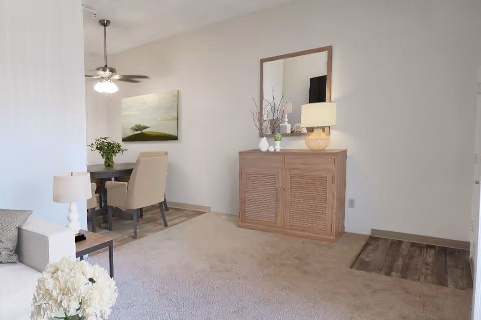 Open living room and dining area with a wooden sideboard and mirror, sofa edge, lamp, and a small dining table with chairs.