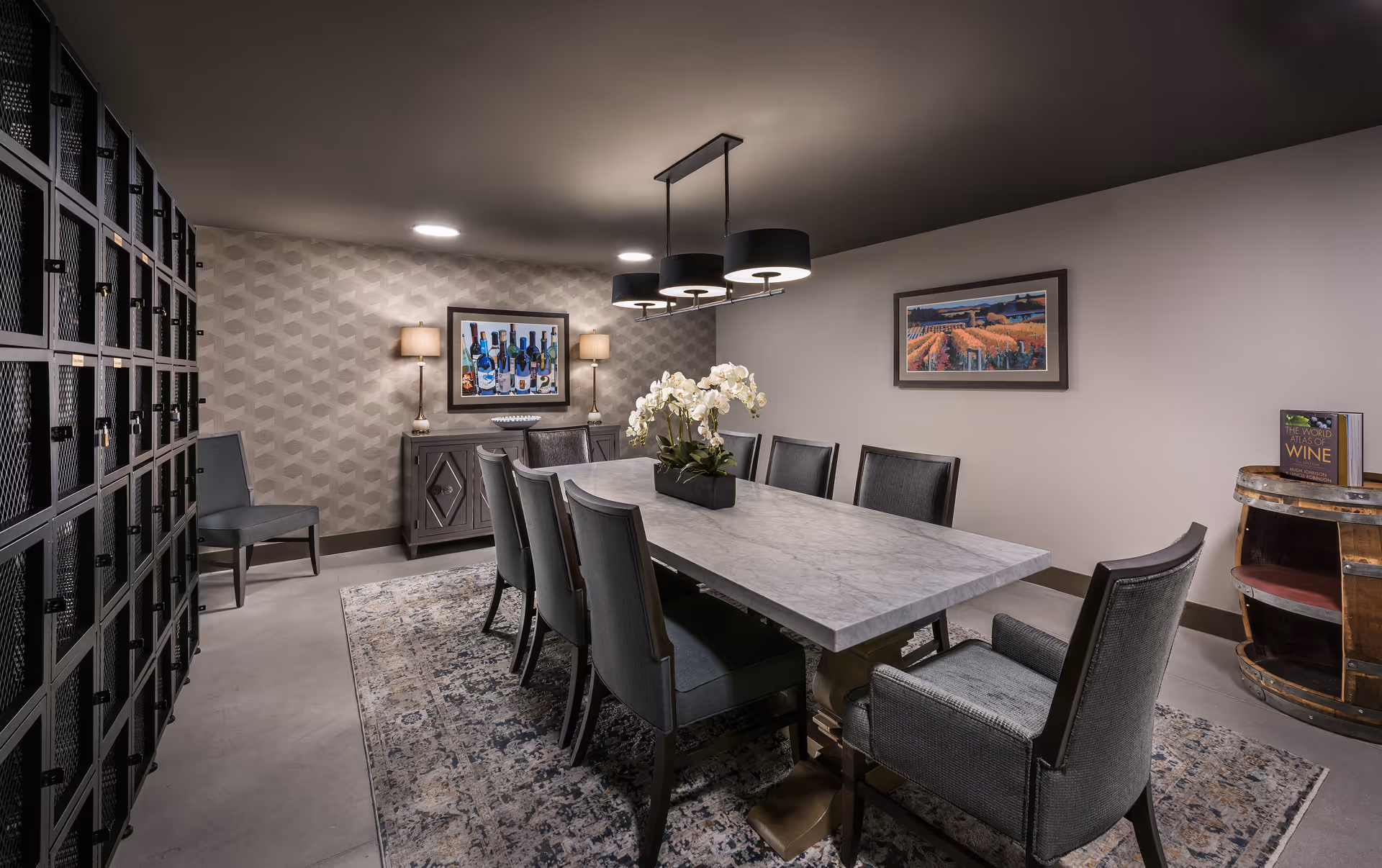 A modern dining room with a long rectangular marble table surrounded by eight upholstered chairs. The room features a patterned area rug, two framed paintings on the walls, a sideboard with two lamps, and a decorative barrel with a book on top. The lighting includes ceiling-mounted fixtures and a three-light pendant above the table. On the left side, there is a wall of black metal lockers.
