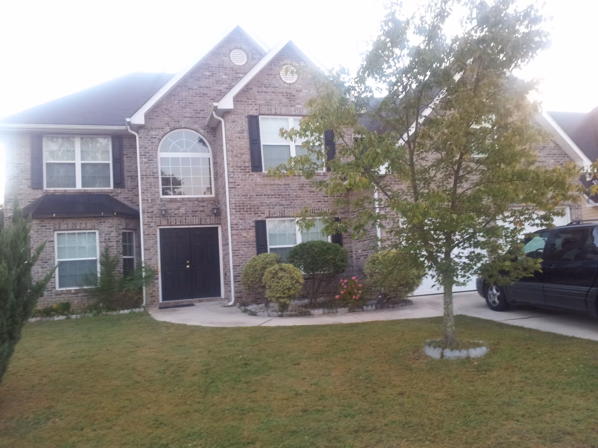 Front exterior view of a two-story brick house with black shutters and a black front door. There is a large arched window above the door, a tree and bushes in the front yard, and a black vehicle parked in the driveway.
