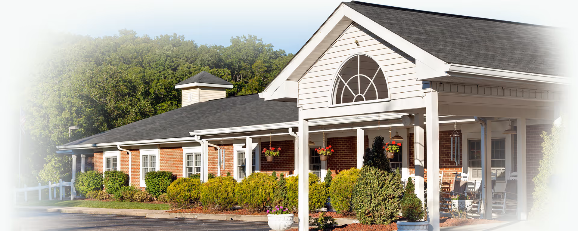 Exterior view of a single-story brick building with a covered porch, white columns, and hanging flower baskets. The building is surrounded by well-maintained shrubs and trees, with a paved driveway in front.