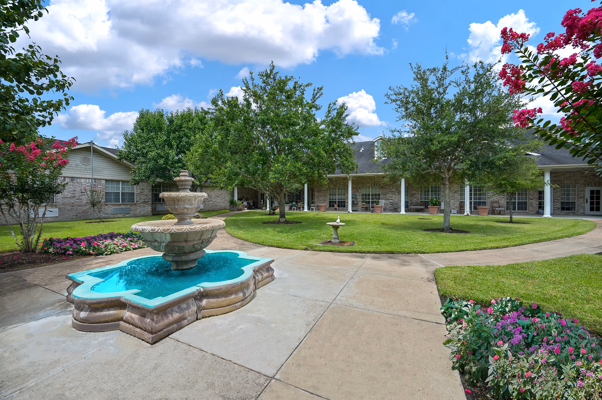 Courtyard with a decorative tiered fountain, paved walkways, manicured lawn, trees, and a covered porch with windows and potted plants in the background.