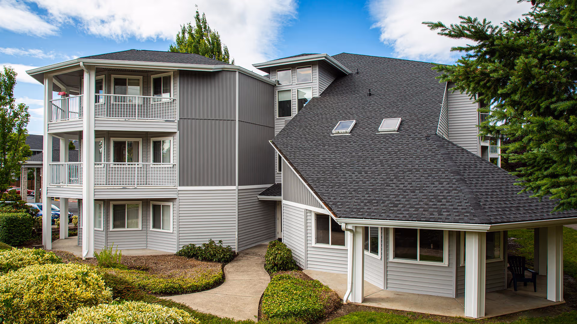 Exterior view of a multi-story senior living facility building with gray siding and white trim. The building features balconies with railings, multiple windows, a sloped roof with skylights, and surrounding greenery including bushes and trees. A concrete pathway curves through the landscaped area in front of the building.