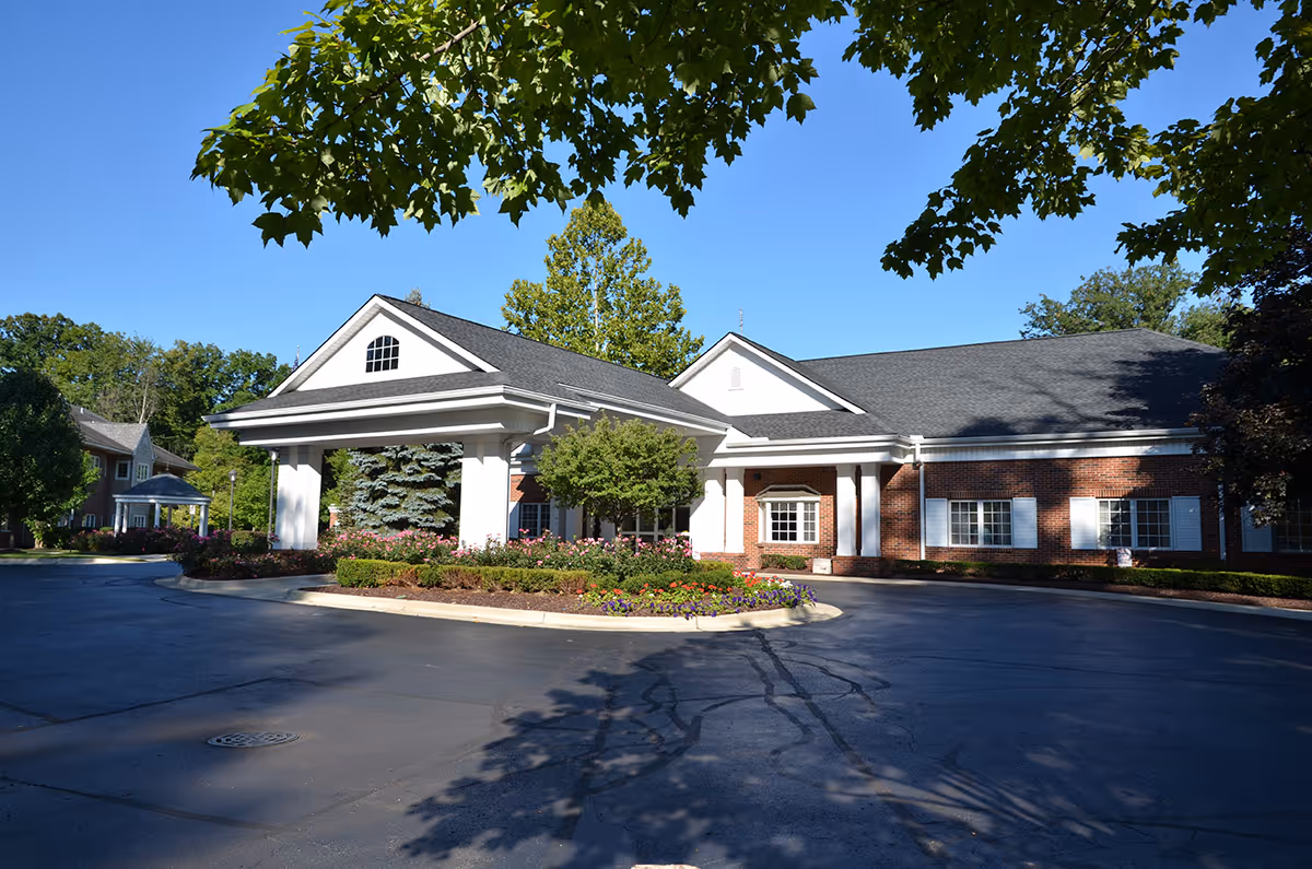 Exterior view of a single-story brick building with white columns and a covered entrance, surrounded by landscaped greenery and trees under a clear blue sky.