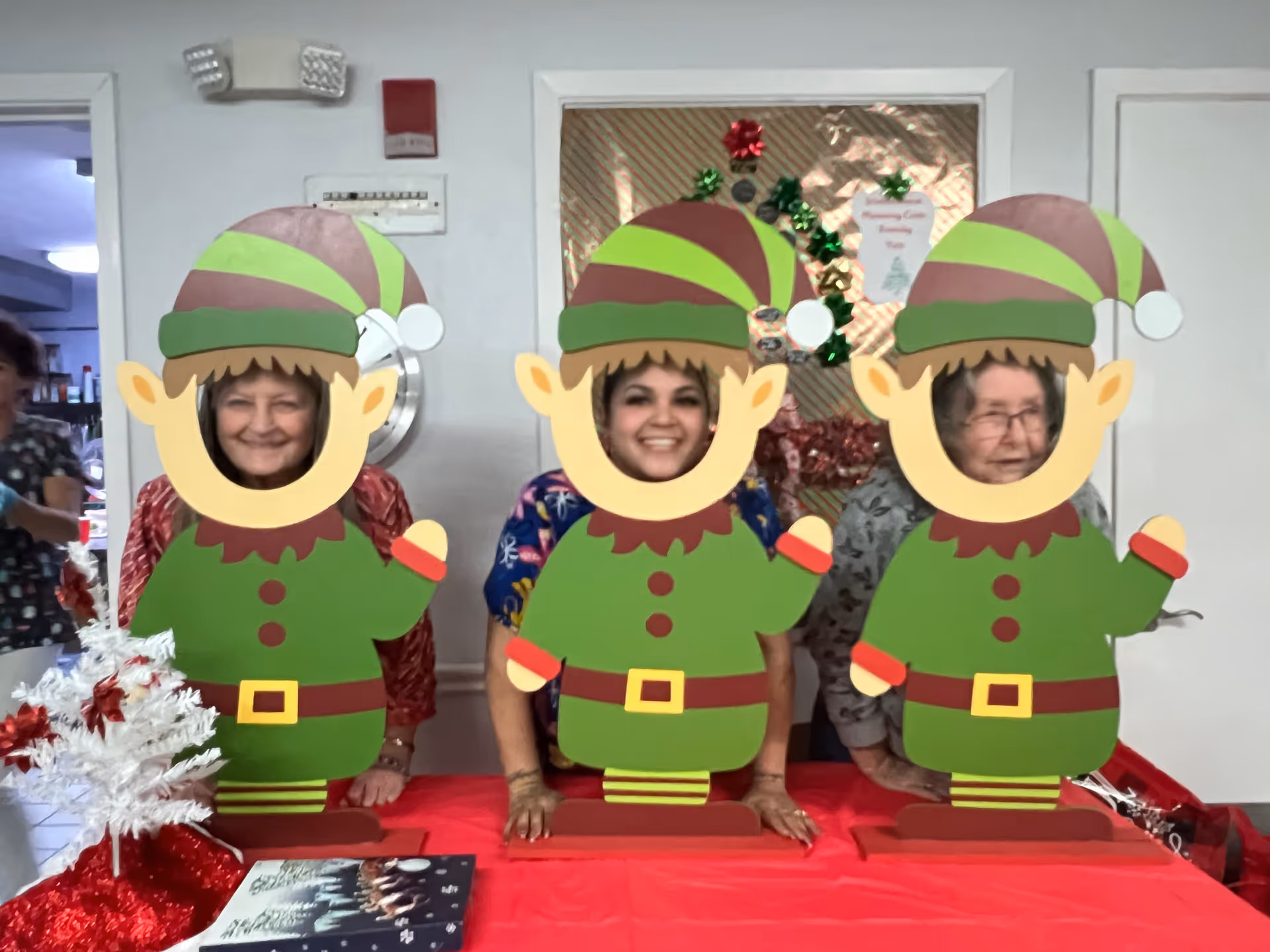 Three elderly women posing behind cutouts of cartoon elves with their faces showing through the holes. They are standing behind a red table with holiday decorations, including a small white Christmas tree with red ornaments. The background shows a decorated window with Christmas-themed items.