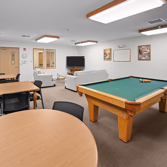 A recreational room featuring a pool table with balls arranged on it, several round and rectangular tables with black chairs, and a seating area with white sofas facing a flat-screen TV mounted on the wall. The room has beige walls, carpeted floor, and ceiling lights with wooden accents. There are framed pictures on the walls and a door on the left side.