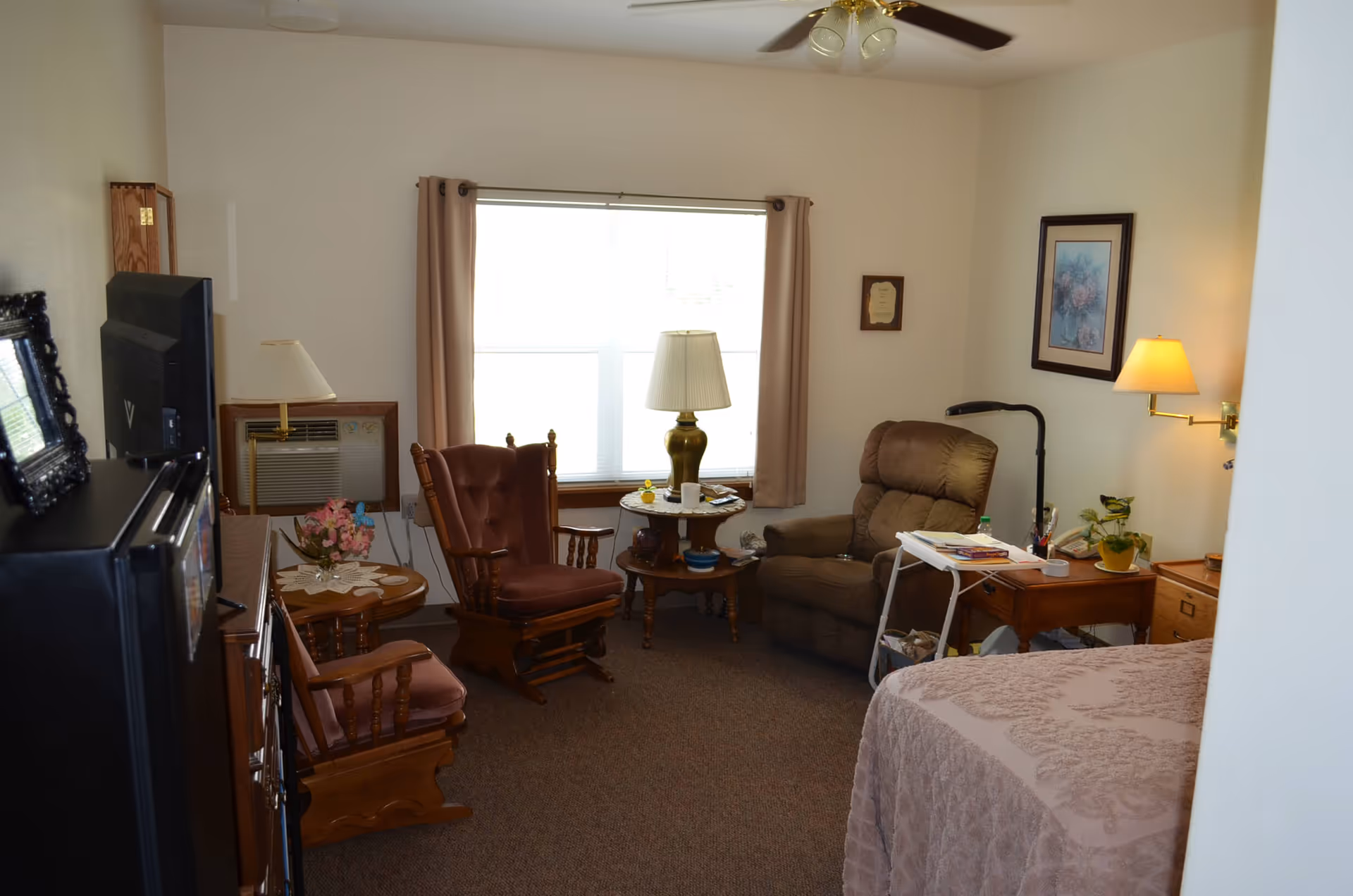Small furnished senior living room with a recliner, wooden rocking chairs, side tables, lamps, and a partially visible bed.