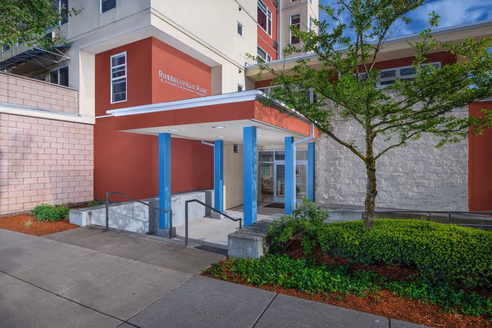 Covered entrance to the Russellville Park building with blue support columns, glass doors, and landscaped greenery.