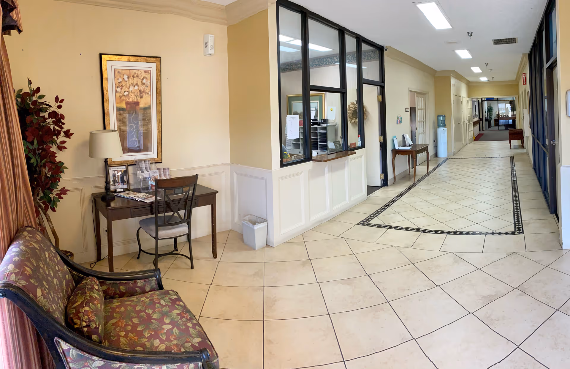 Tiled lobby hallway with a small desk and seating area beside a glass-front reception window.