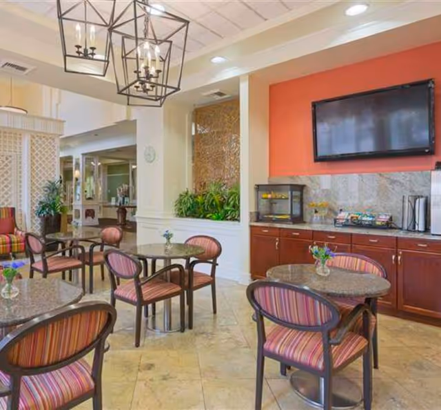 A bright common area with several round tables and striped cushioned chairs. The room features a countertop with a small food display case and a mounted flat-screen TV on a red accent wall. Decorative hanging light fixtures and indoor plants add to the welcoming atmosphere.
