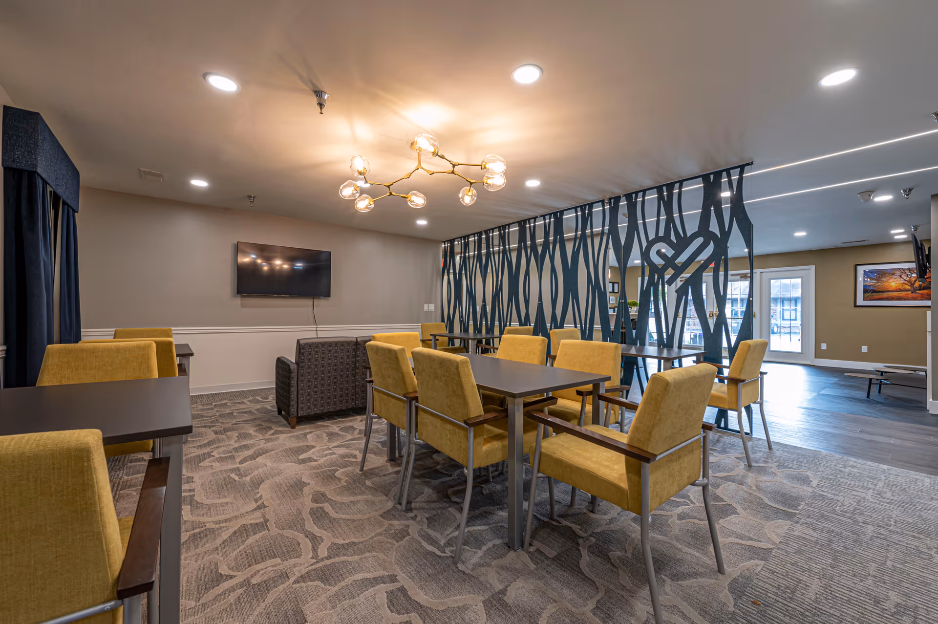 Interior view of a senior living facility common area with multiple tables and yellow upholstered chairs arranged on a patterned carpet. A modern chandelier hangs from the ceiling, and a decorative partition with a heart design separates the space. A wall-mounted TV and a gray armchair are visible in the background, along with large windows and a door letting in natural light.