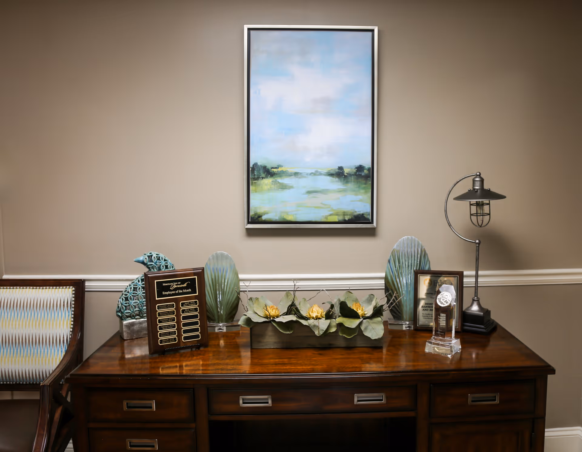 A wooden desk with decorative items including a lamp, plaques, a flower arrangement, and two green ornamental sculptures. Above the desk hangs a framed painting of a serene landscape with a blue sky and greenery. A patterned chair is partially visible on the left side against a beige wall with white trim.