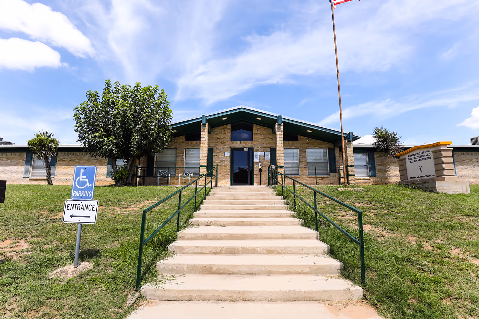 Front entrance of a single-story brick rehabilitation and healthcare center with steps and handrails leading up, a flagpole, landscaping, and a handicapped parking/entrance sign.