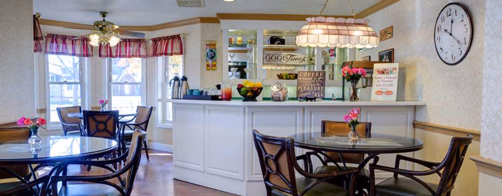 A cozy dining area with round glass-top tables and wooden chairs, each table decorated with a small vase of pink flowers. There is a white counter with various decorative signs and items, including a bowl of fruit and a coffee-themed sign. The room has large windows with red valance curtains, a ceiling fan with lights, and a large wall clock.