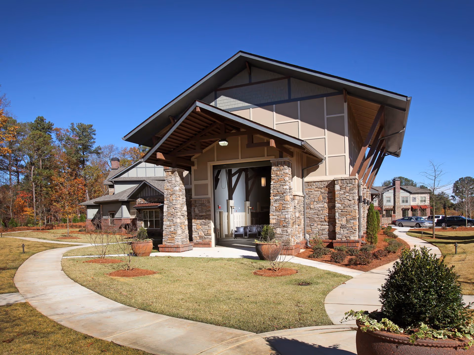 Front entrance of a modern senior living building with stone columns, a covered porte-cochere and curved walkways through landscaped grounds.