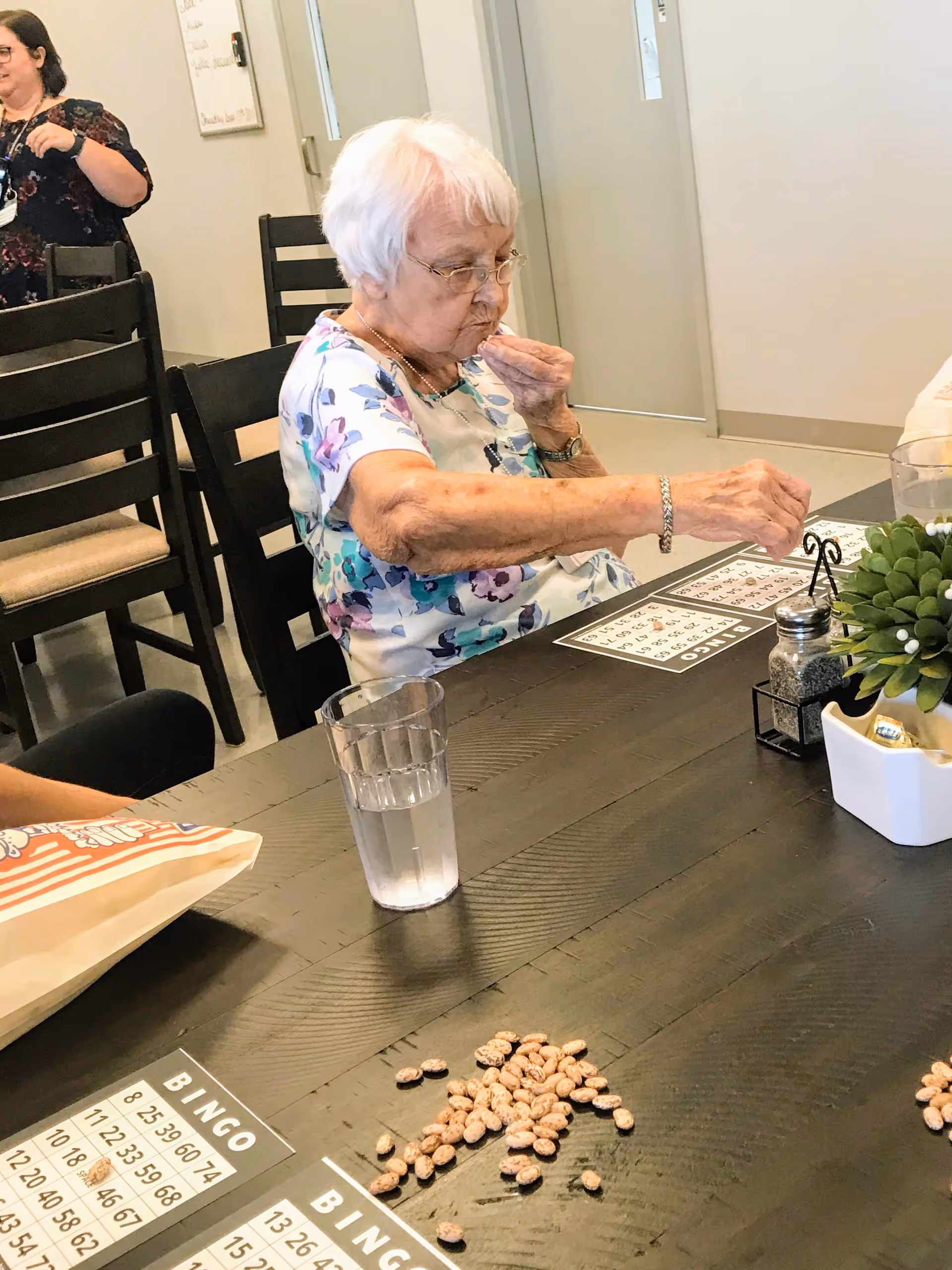An elderly woman seated at a table in a communal room playing bingo and placing beans on a bingo card.