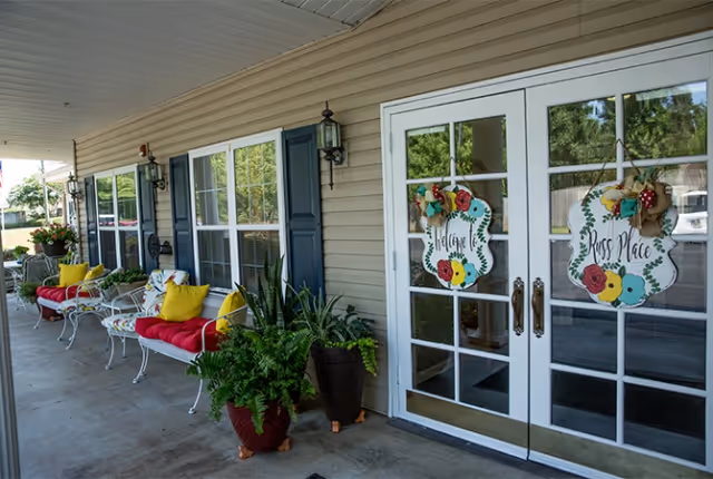 Covered porch area at Trustwell Living at Russ Place with white-framed glass double doors decorated with floral welcome signs. The porch has beige siding, black shutters, outdoor wall lanterns, white metal benches with red cushions and yellow pillows, and potted green plants.
