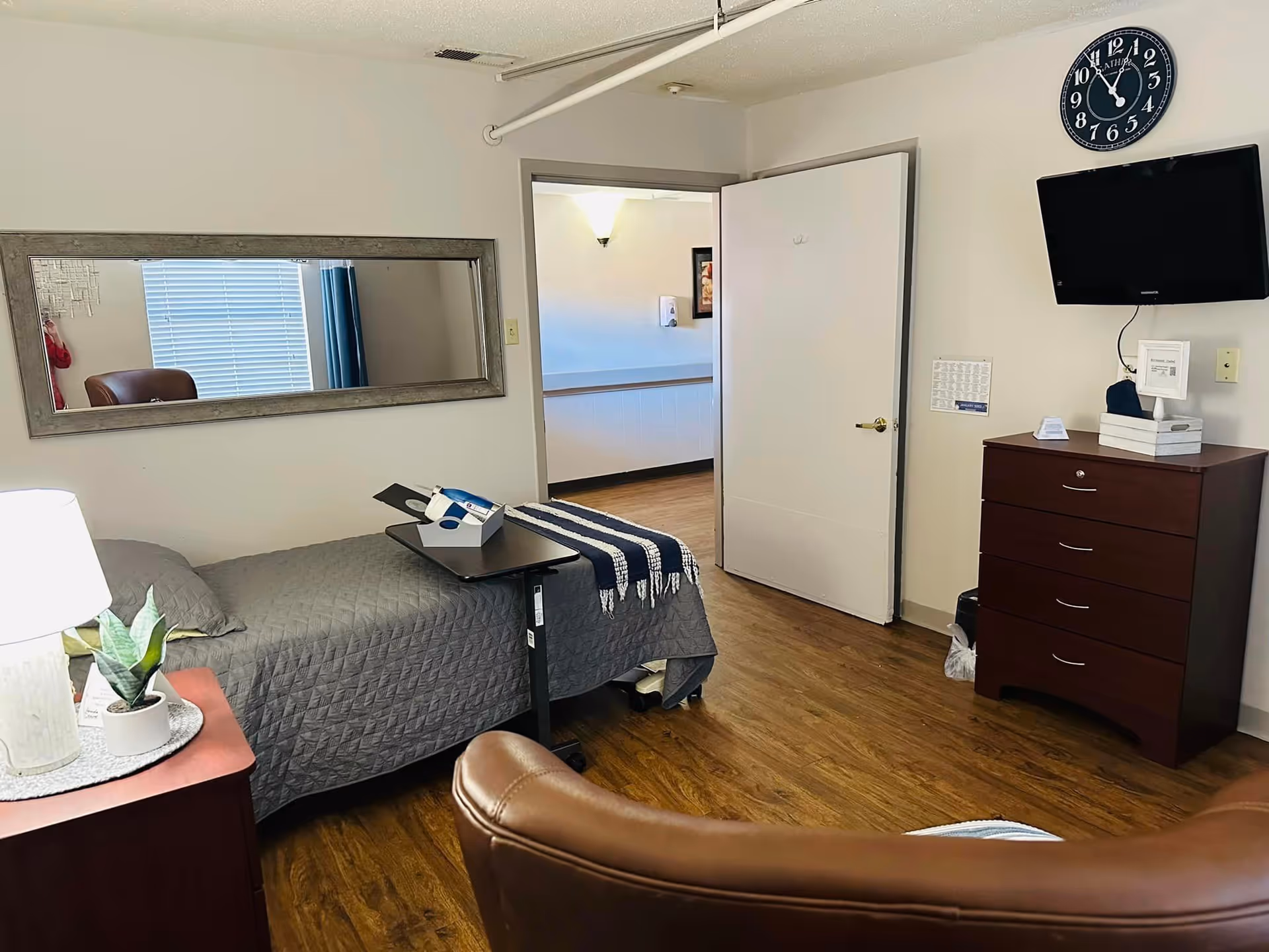 A tidy nursing facility bedroom with a single bed, bedside table and lamp, overbed tray, dresser with a wall-mounted TV and clock, and an open door to a hallway.