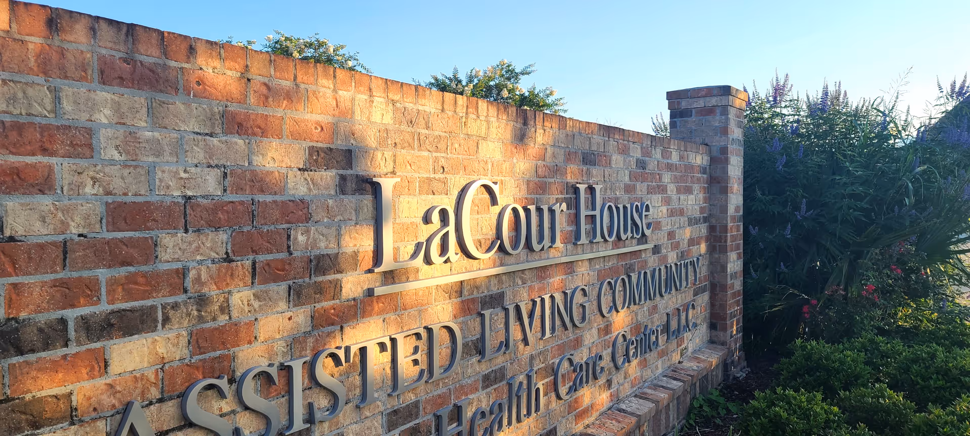A brick wall sign for LaCour House Assisted Living Community with surrounding greenery and flowers under a clear blue sky.