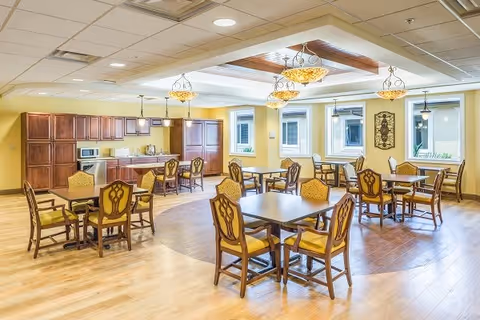 A bright and spacious dining area in a senior living facility with several wooden tables and chairs arranged neatly. The room features a kitchen area with wooden cabinets, a microwave, and a refrigerator. The ceiling has decorative light fixtures and a recessed design with wood accents. Large windows allow natural light to fill the space.