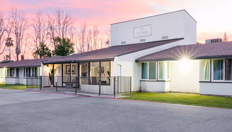 Exterior view of San Joaquin Nursing and Rehabilitation Center building at sunset with a clear sky and leafless trees in the background. The building is single-story with a white facade, brown roof, and several windows. There is a fenced porch area with some plants and framed pictures visible inside.