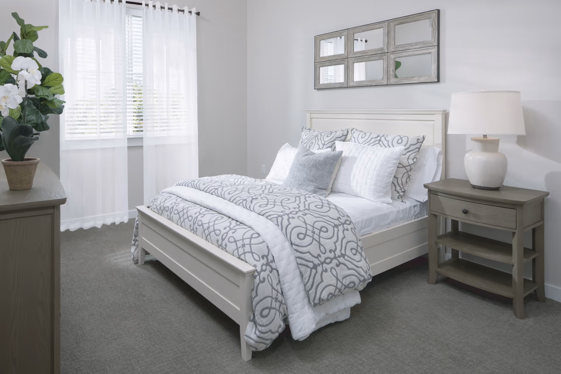 Bright, neatly made bedroom with a white bed, patterned bedding, nightstand with a lamp, dresser, and a window with sheer curtains.
