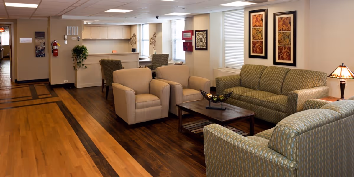 A cozy living room area in a senior living facility with two beige armchairs, two patterned green sofas, a wooden coffee table with a decorative bowl, and a side table with a lamp. The room has wooden floors, light-colored walls, framed artwork, and large windows with blinds allowing natural light.