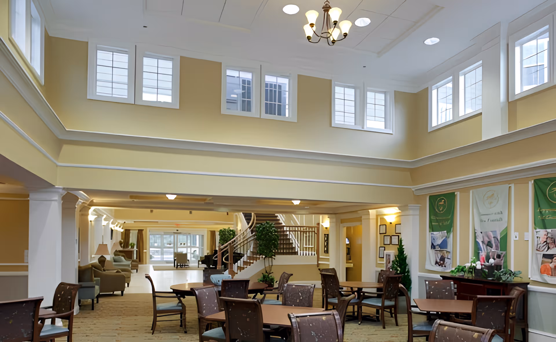 Spacious two-story senior living common room with dining tables and chairs, seating areas, a central staircase, and high windows.