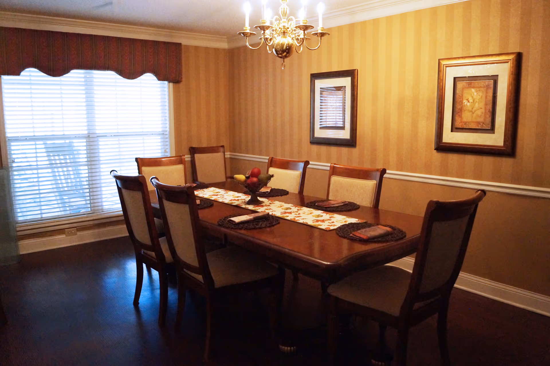 A dining room with a wooden table set for six people. The table has a decorative runner and placemats with folded napkins. There is a chandelier hanging above the table, two framed pictures on the striped wallpapered wall, and a large window with blinds and a valance.