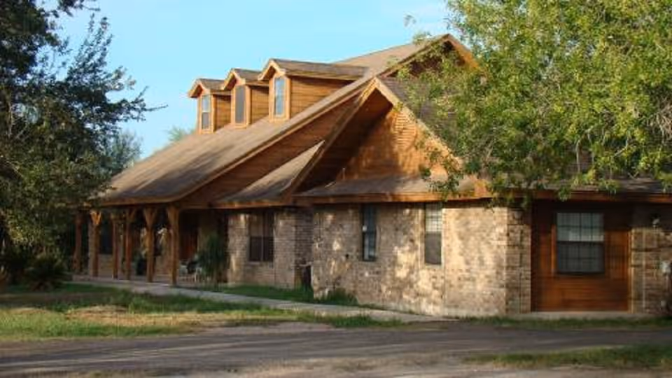Exterior view of a single-story building with a combination of brick and wood siding, featuring a sloped roof with three dormer windows. The building is surrounded by trees and greenery, with a paved driveway or road in front.