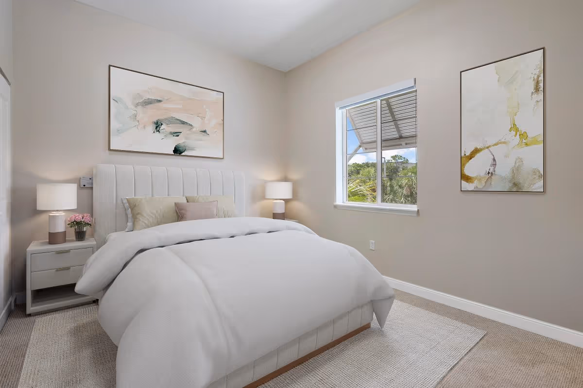 A neatly made bed with a white upholstered headboard in a bedroom. The bed has white bedding and several pillows. There are two bedside tables with matching lamps on each side of the bed. The walls are painted beige and decorated with two abstract paintings. A window shows greenery outside.