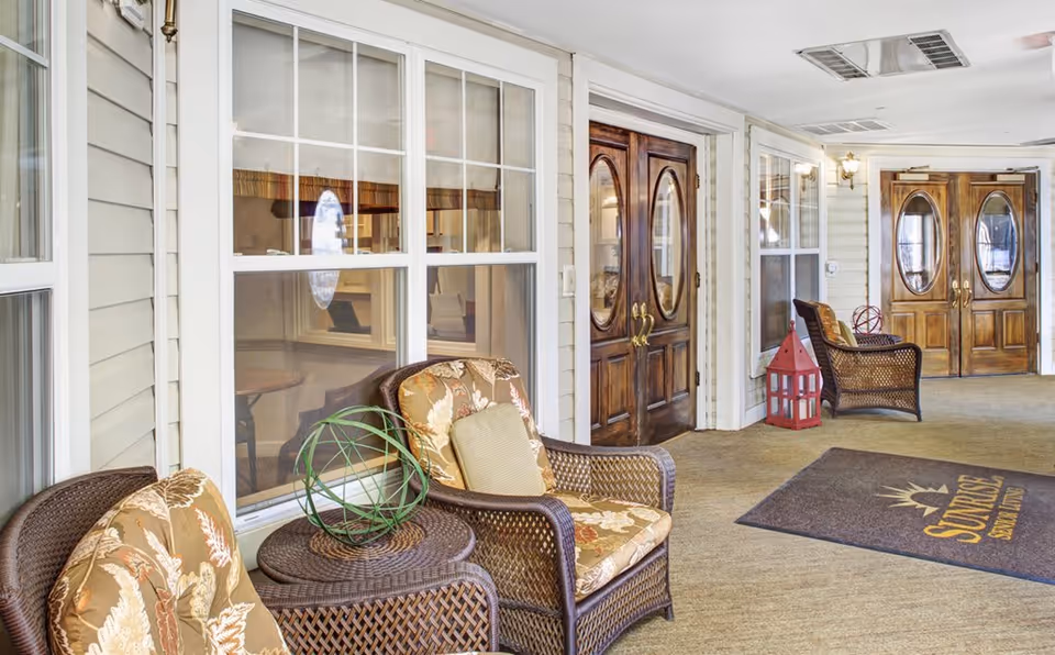 A covered outdoor seating area at Sunrise of Granite Run featuring wicker chairs with floral cushions, a small wicker table with a decorative green orb, large wooden double doors with oval glass panels, and a floor mat with the Sunrise logo.
