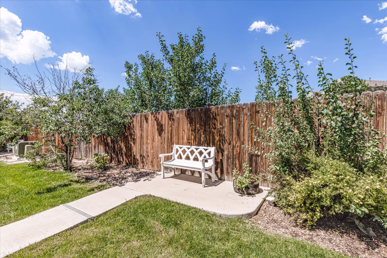 Outdoor garden area with a white bench on a concrete slab, surrounded by green grass, plants, and trees, with a wooden fence in the background under a blue sky with some clouds.