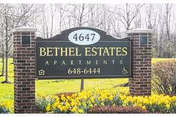 A brick signpost with a black and gold sign reading '4647 Bethel Estates Apartments 648-6444' surrounded by yellow flowers and grass with leafless trees in the background.