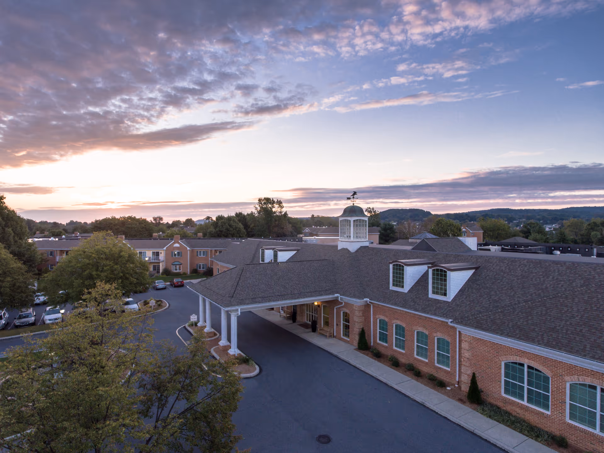 Exterior view of The Highlands at Wyomissing senior living facility during sunset, showing a large brick building with a covered entrance, multiple windows, and a cupola on the roof. Trees and parked cars are visible around the building with a partly cloudy sky in the background.