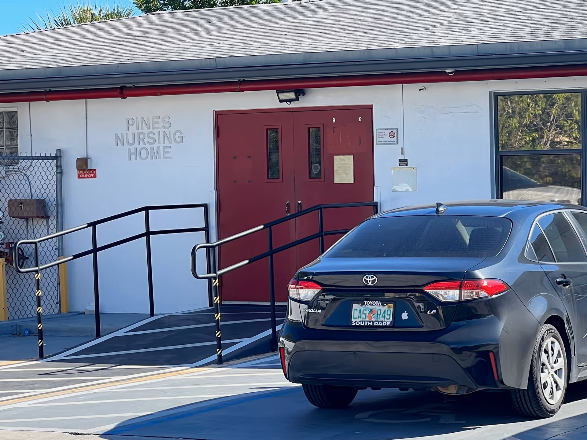 Exterior view of Pines Nursing Home showing a white building with a gray roof, red double doors, a wheelchair ramp with black railings, and a black Toyota Corolla parked in front with a South Dade license plate.