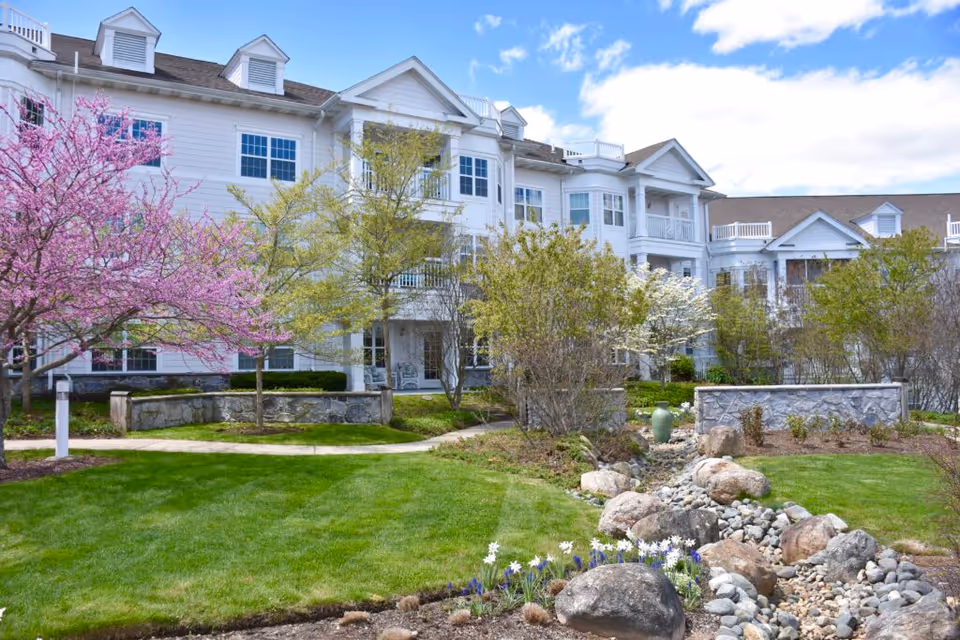Exterior view of a senior living facility with white multi-story buildings, surrounded by a landscaped garden featuring green grass, blooming trees with pink and white flowers, shrubs, rocks, and a small dry creek bed under a partly cloudy blue sky.