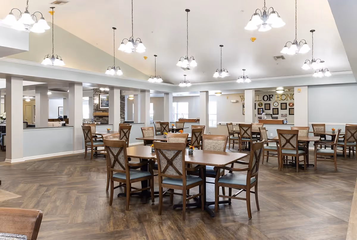 A spacious dining room with multiple wooden tables and chairs arranged neatly. The room features a high ceiling with several hanging light fixtures, wood-patterned flooring, and a wall in the background with a 'Wall of Honor' display. The area appears clean and well-lit with natural light coming through the windows.