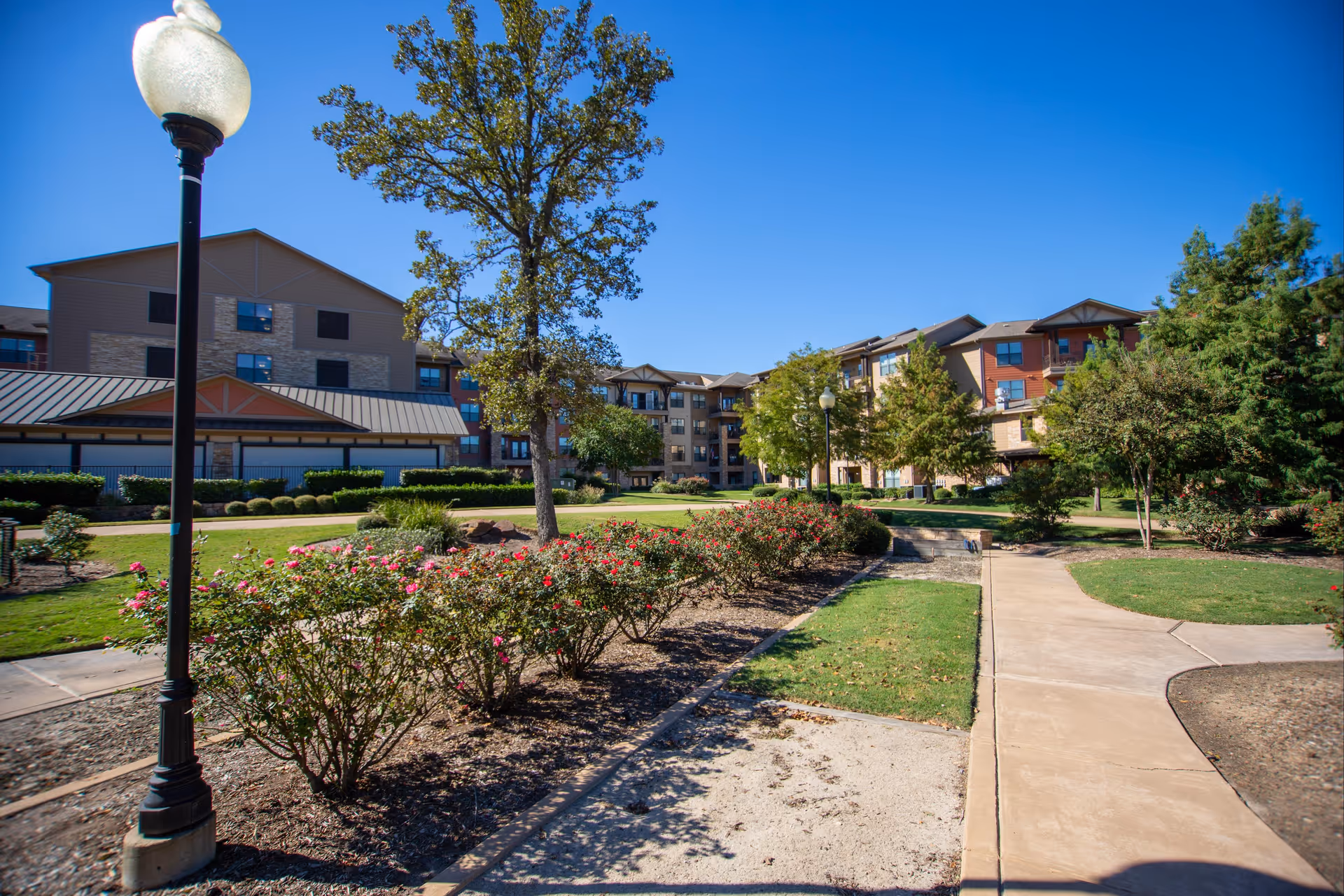 Outdoor garden area with a paved walkway, flower bushes, trees, and a lamppost in front of a multi-story residential building under a clear blue sky.