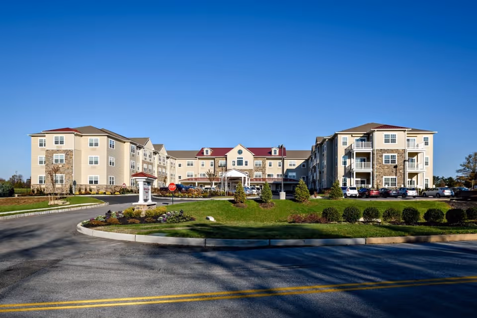 Exterior front view of a large three-story senior living facility building with beige siding and stone accents, a red roof, multiple windows, balconies, a driveway with a stop sign, landscaped greenery, and parked cars under a clear blue sky.