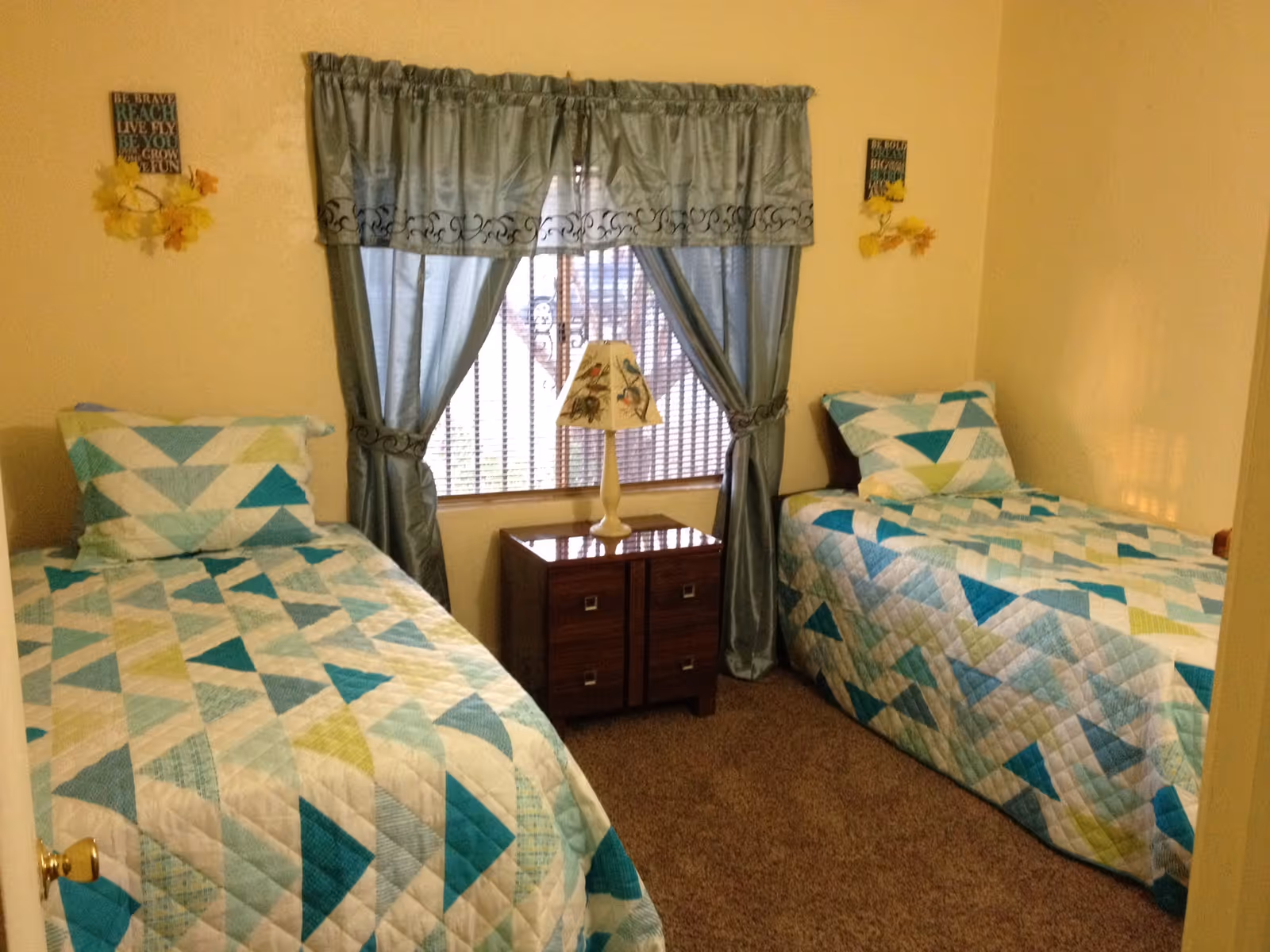 A small bedroom with two twin beds placed on opposite sides of the room. Each bed has a quilted bedspread with a geometric pattern in shades of blue, green, and white. Between the beds is a wooden nightstand with two drawers, topped with a decorative lamp featuring a bird design. A window with blue curtains tied back is centered on the wall behind the nightstand. The walls are painted a light beige color and have small decorative plaques with inspirational quotes and artificial yellow flowers.