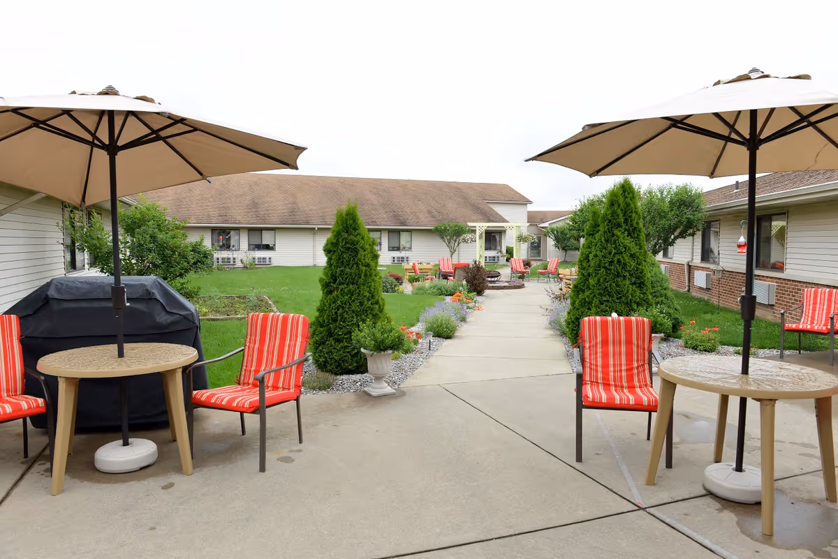 Outdoor courtyard area at The Laurels of Defiance featuring a concrete walkway lined with green shrubs and colorful flowers. There are multiple red and white striped cushioned chairs around beige tables with large beige umbrellas providing shade. The surrounding buildings have beige siding and windows with air conditioning units.