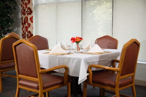 A dining table set for four with white tablecloth, folded white napkins, placemats, wine glasses, and a small vase with red flowers in the center. The table is surrounded by four brown cushioned chairs with wooden frames. The background shows large windows with closed blinds and patterned curtains on the side.