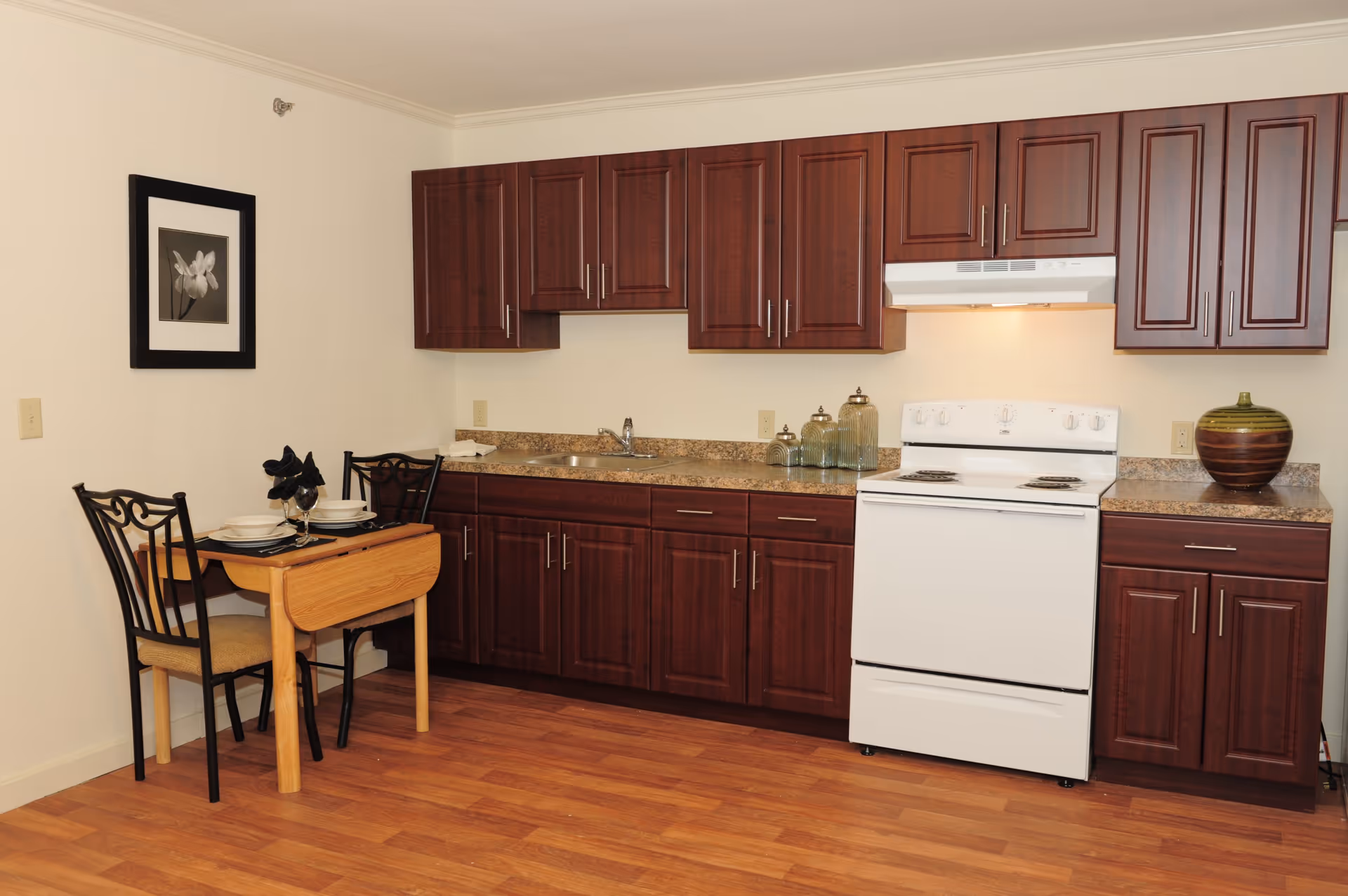 Compact kitchen with dark wood cabinets, a white stove and sink, and a small dining table set against a light wall.
