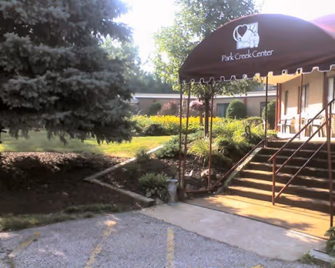 Exterior view of Park Creek Center showing an entrance with a maroon awning displaying the facility's name and logo. There are stairs leading up to the entrance, surrounded by landscaped greenery and trees.