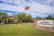 Exterior view of Opp Health and Rehabilitation facility with a large green lawn, an American flag on a flagpole, trees, and a clear sky with scattered clouds. The building has a covered entrance and a sign that reads 'Opp Health and Rehabilitation'.