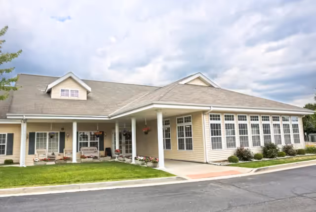 Single-story beige senior living building with a covered entrance, large windows, a small landscaped lawn, and a cloudy sky above.