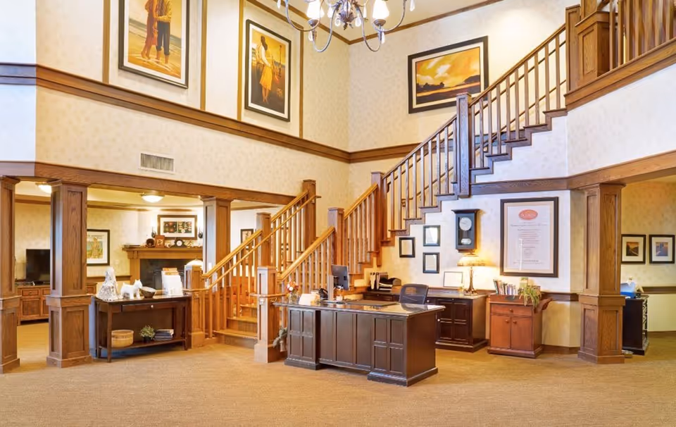 Interior view of a senior living facility lobby with a wooden reception desk, staircase with wooden railings, framed artwork on the walls, and warm lighting. There is a seating area visible through an open doorway with a fireplace and TV.