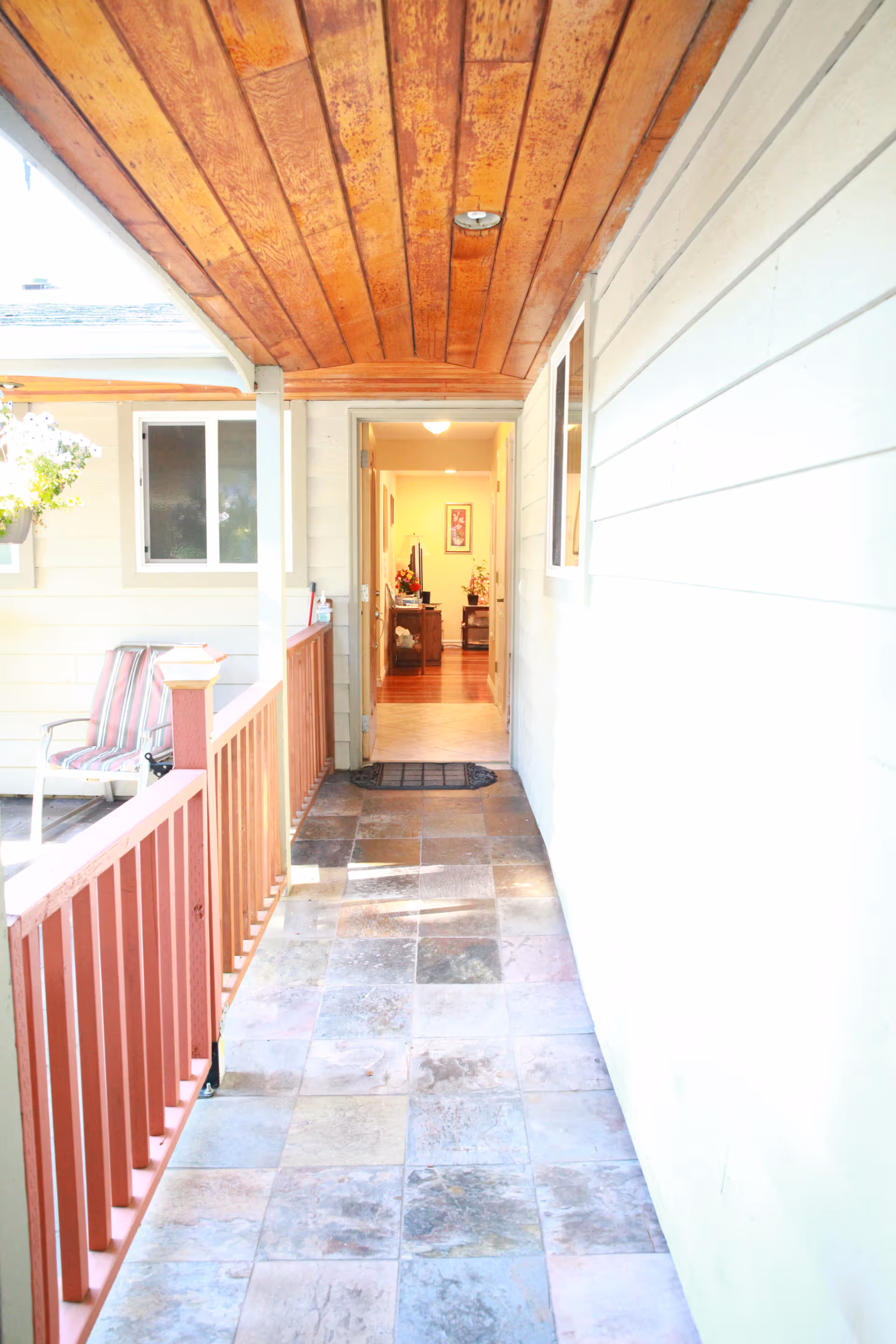 A covered outdoor walkway with tiled flooring and a wooden ceiling leading to an open door into an interior space. To the left, there is a red railing and a striped chair next to a window. The interior visible through the door shows a hallway with wooden flooring and furniture.